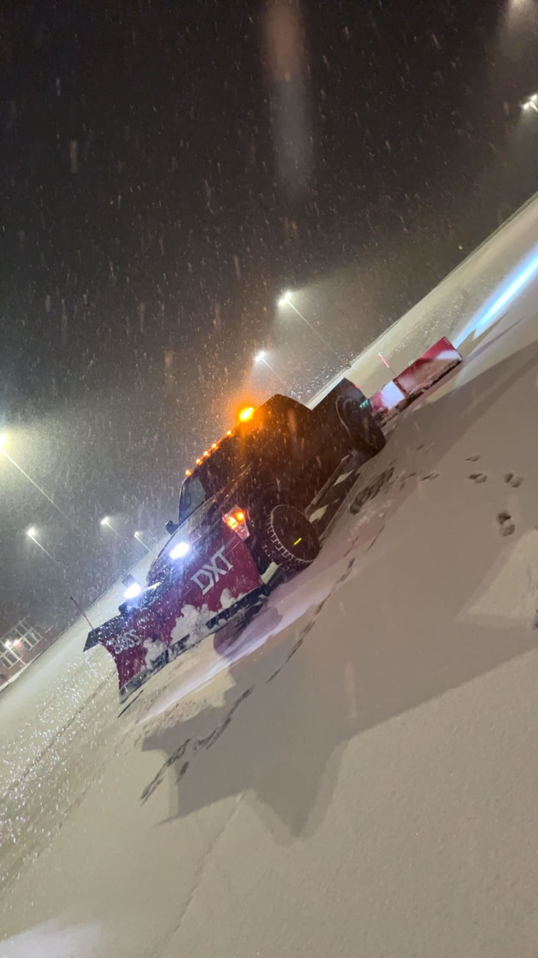 Truck with plow clearing snow at night in a snowy parking lot during a snowstorm.