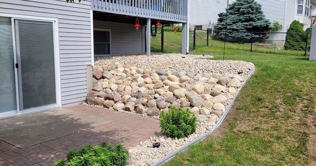 Landscaped backyard featuring stone wall, gravel area, and green plants near patio.