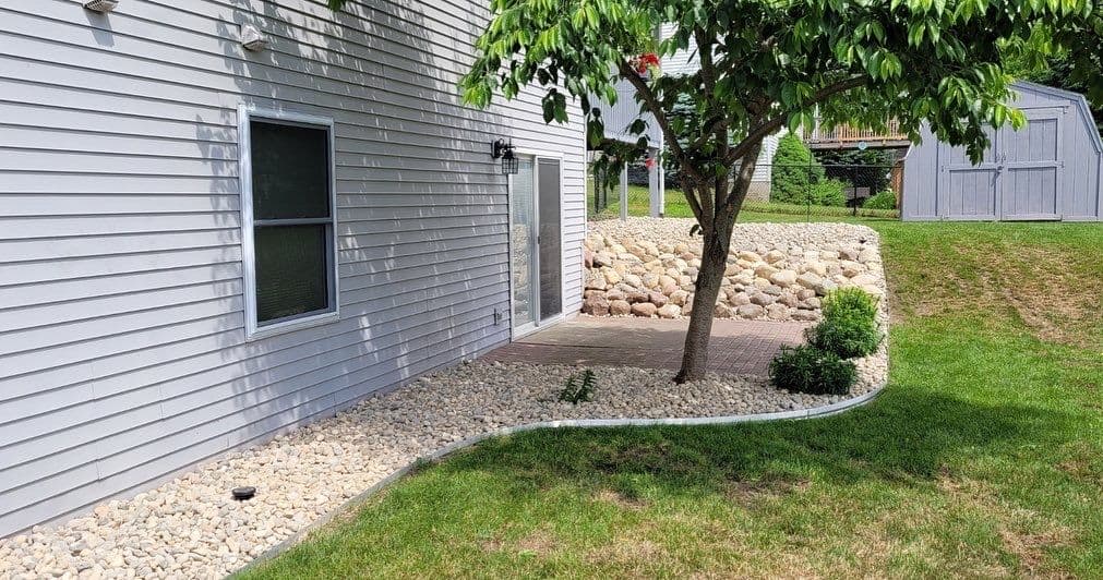 Landscaped backyard featuring a tree, stone border, and patio area beside a house.