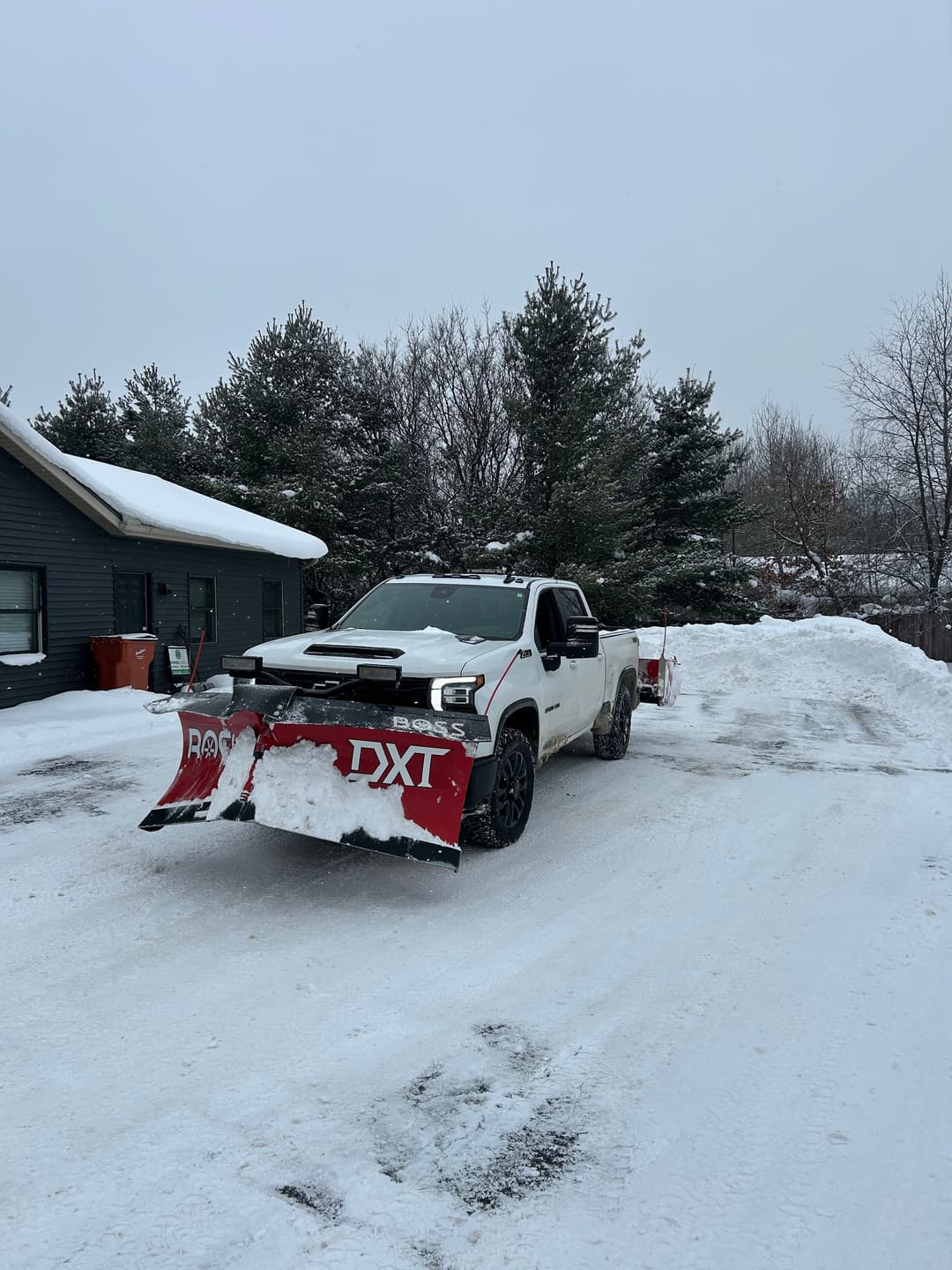 White pickup truck with snow plow clearing driveway in snowy residential area.