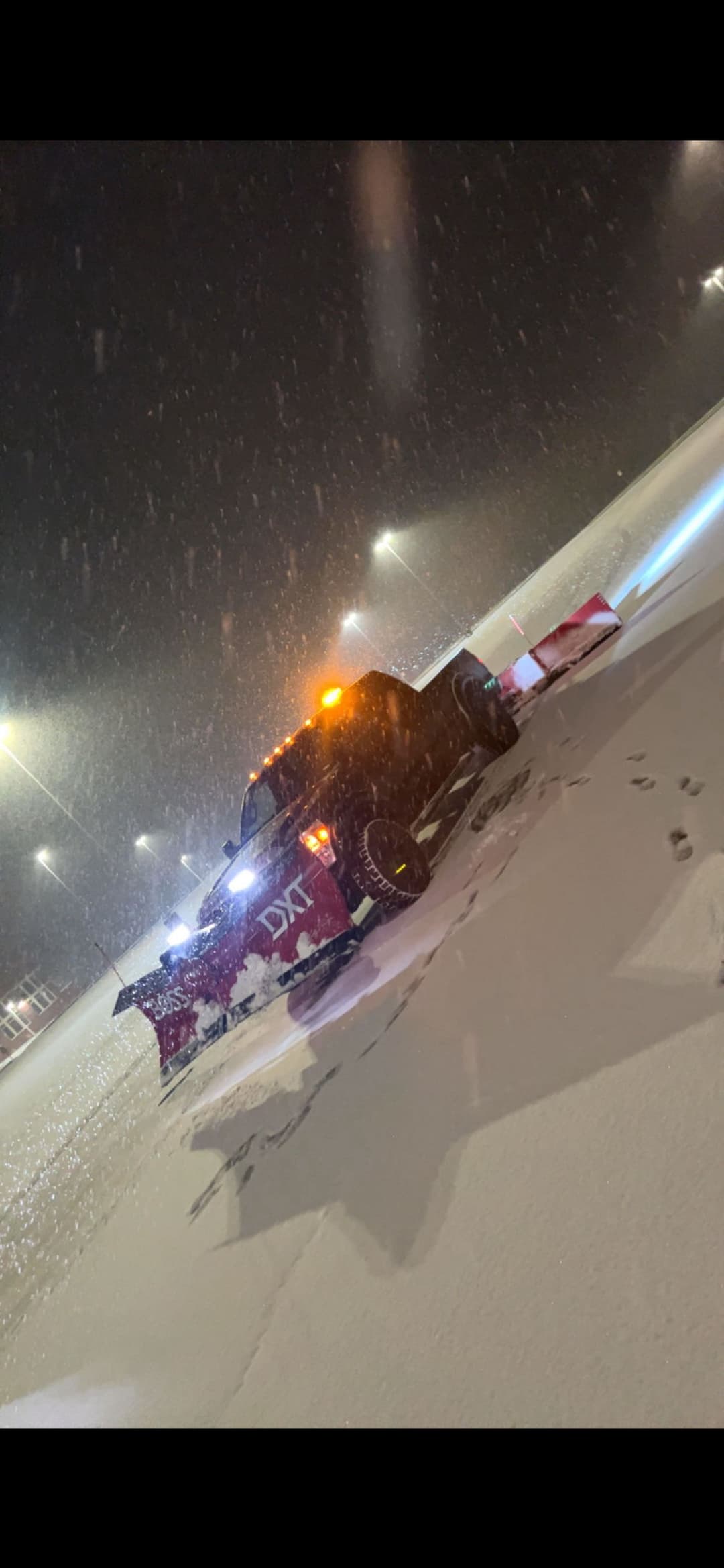 Snowplow truck clearing snow at night with heavy snowfall and bright lights.