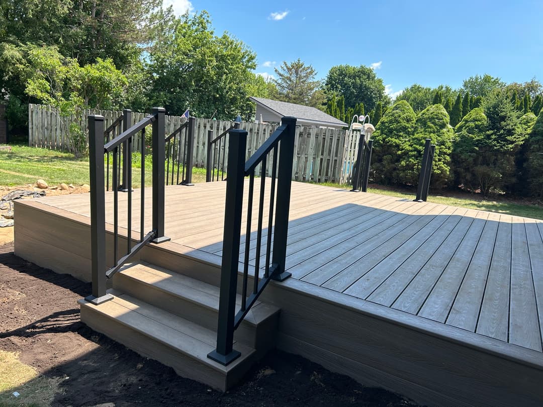 Newly built wooden deck with black railing and stairs, surrounded by green landscaping.