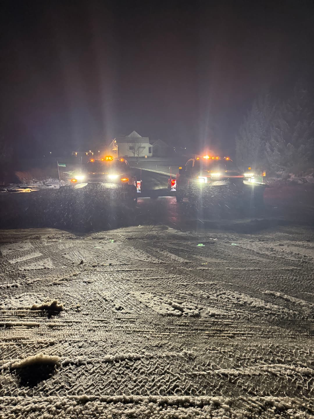 Two snow plows working at night on a snowy road, illuminated by bright lights.