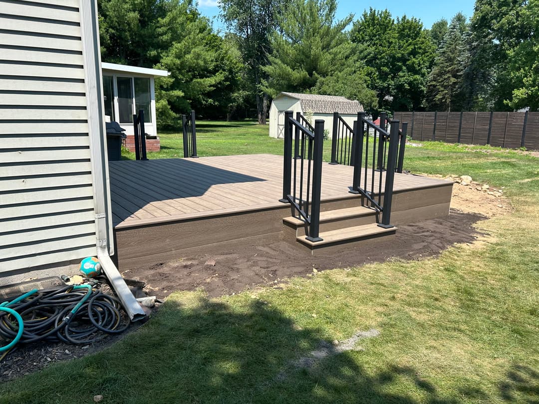 New wooden deck with black railing and stairs, surrounded by green grass and trees.