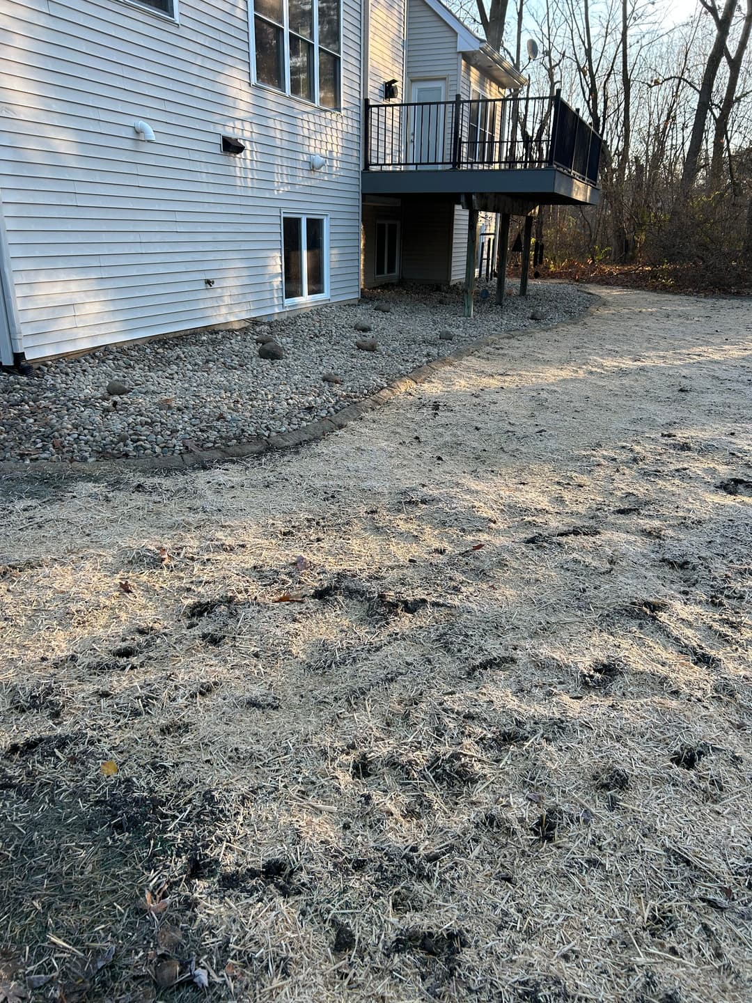 Backyard landscape showing freshly laid straw, gravel path, and a house foundation with deck.